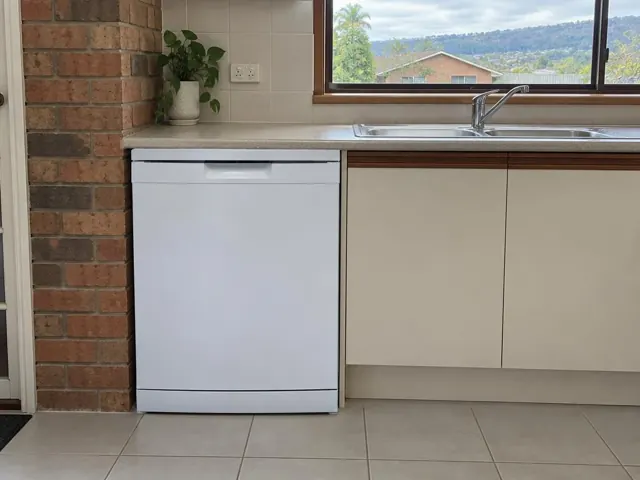 White dishwasher installed under a kitchen benchtop beside a sink, brick wall and tiled kitchen in Tingira Heights